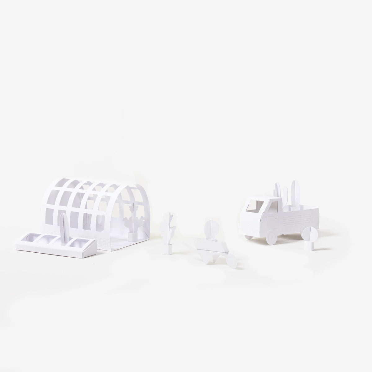 A set of minimalist white paper models, including an arched greenhouse structure, a wheelbarrow, plants and a pickup truck, arranged on a seamless white background.