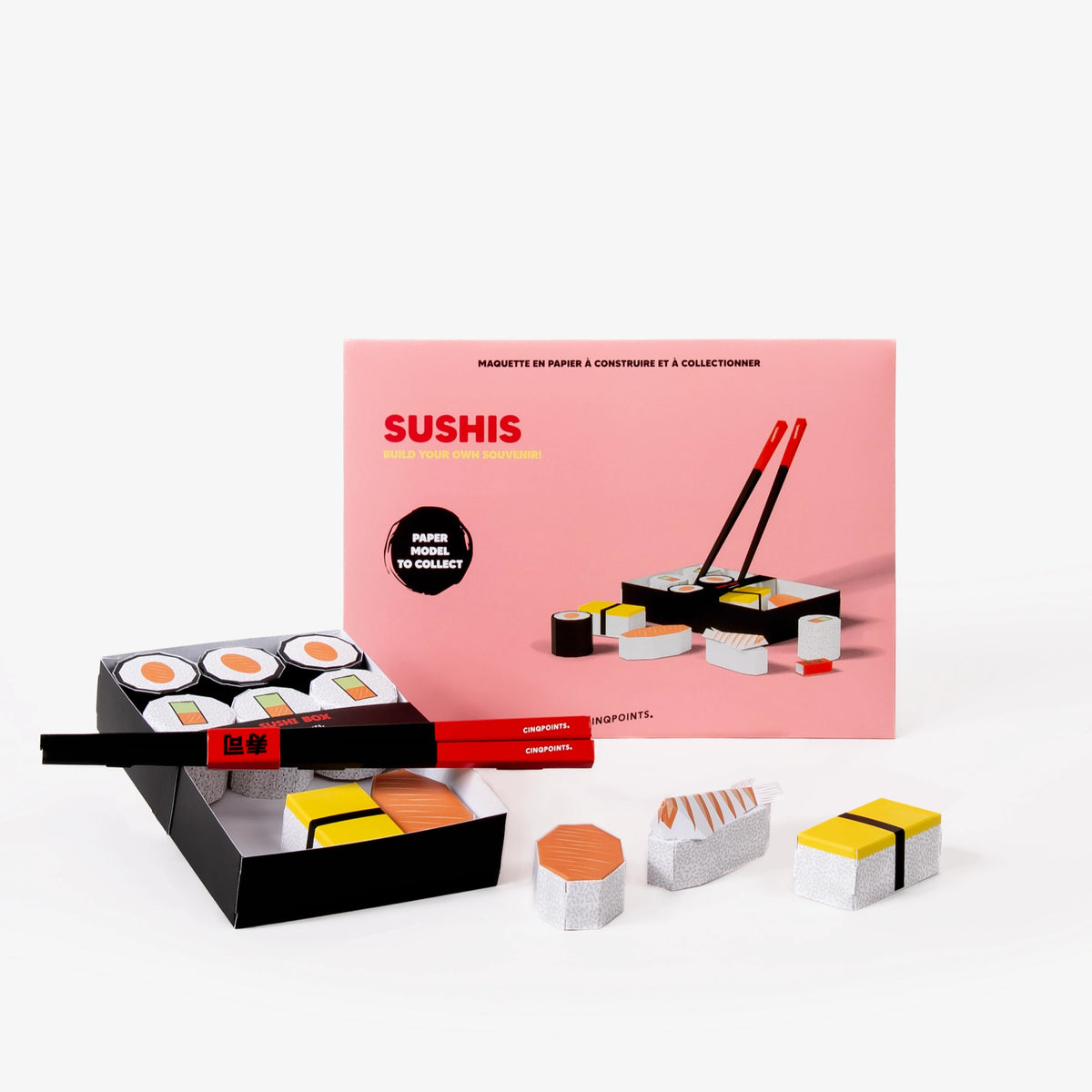 Sushi-themed paper craft kit with a pink envelope behind, with a black tray of paper sushi pieces, red chopsticks, and several small paper sushi models arranged on a white background.