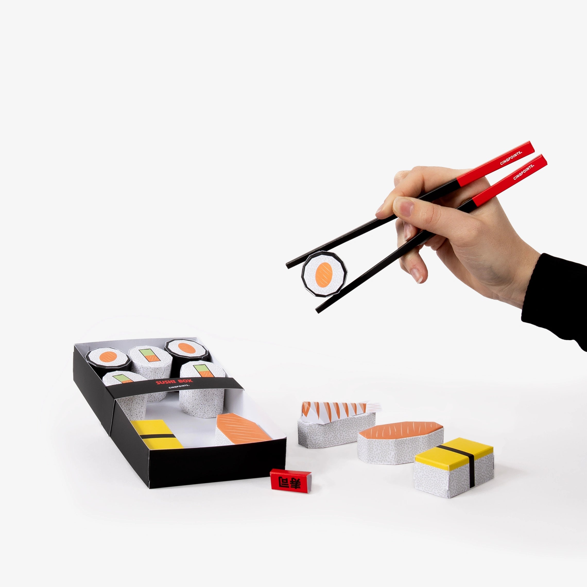 Hand holding paper chopsticks picking up a paper model sushi roll with a box of sushi and additional sushi pieces on a white background.