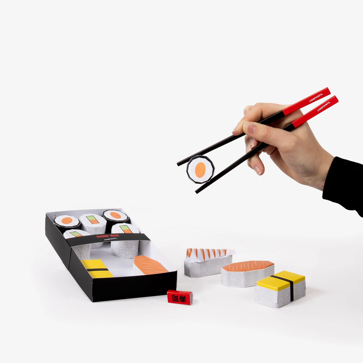 Hand holding paper chopsticks picking up a paper model sushi roll with a box of sushi and additional sushi pieces on a white background.