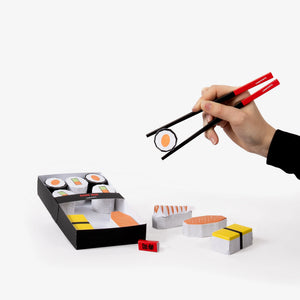 Hand holding paper chopsticks picking up a paper model sushi roll with a box of sushi and additional sushi pieces on a white background.