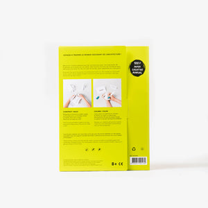 The back cover of a yellow cardboard folder of a paper model kit stands upright on a clean white background, showing instructional text, and two small photos of hands working with white paper.