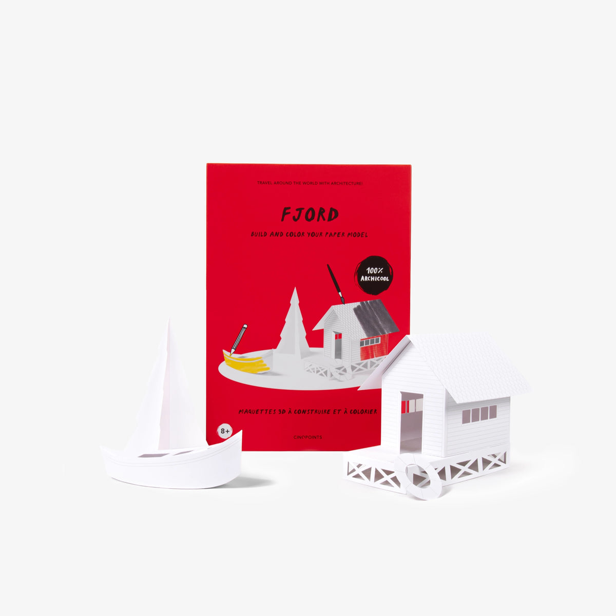 A red cardboard folder of a paper model kit stands upright behind a few white small 3D paper model of a house, boat, and tree on a clean white background.