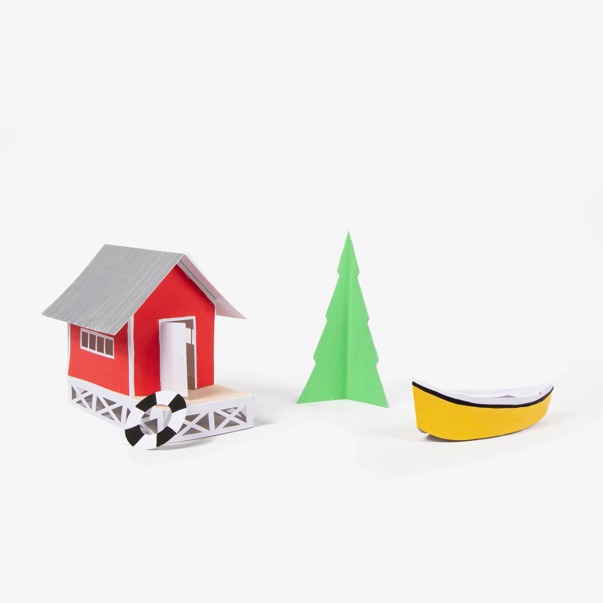 A set of minimalist, geometric paper toy models on a clean white background, including a red house with a gray roof, a green pine tree, and a yellow boat.